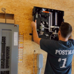 A Postma electrician installing or inspecting an electrical panel, wearing a Postma company shirt in a workshop environment.