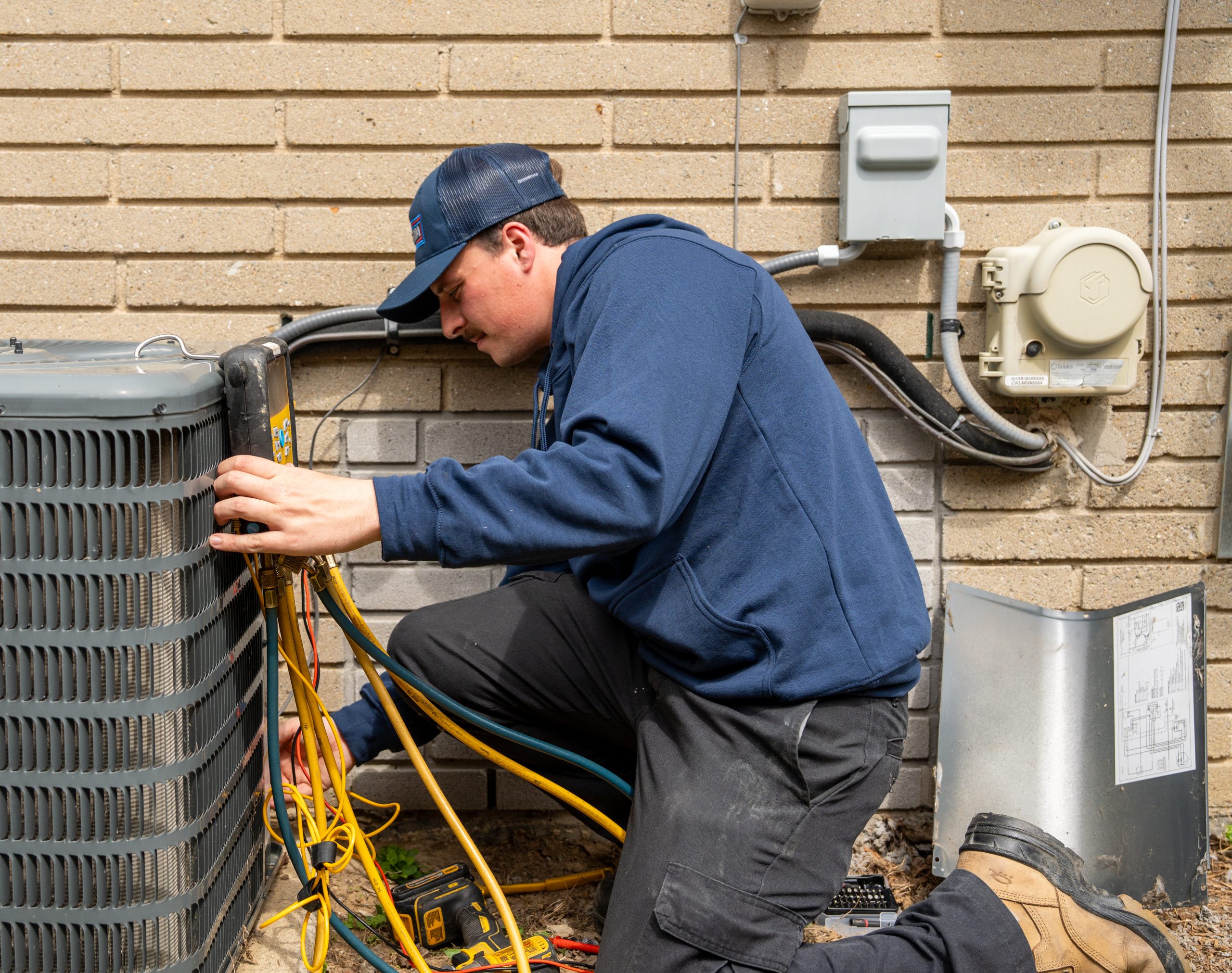 Technician performing air conditioner maintenance, checking refrigerant lines and connections, with tools and equipment visible in a residential setting.