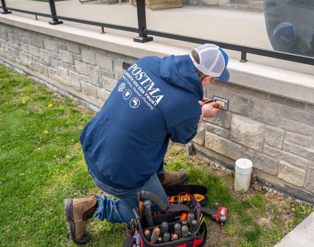Electrician from Postma Heating & Cooling working on outdoor electrical outlet installation, wearing branded hoodie, using tools, surrounded by grass and stone wall.